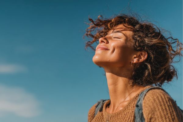 A woman with curly hair smiles gently with eyes closed, face lifted toward the sun against a clear blue sky, conveying a sense of calm and well-being.