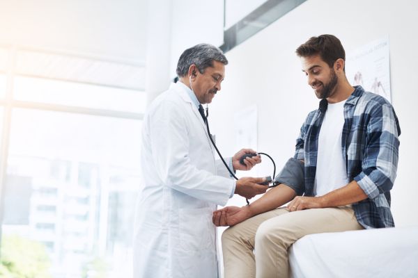 A doctor measures a patient’s blood pressure during a routine checkup in a bright medical exam room, with the patient seated on an exam table and smiling.