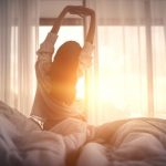 A woman sits on a bed stretching her arms overhead as warm morning sunlight streams through sheer curtains, symbolizing refreshed sleep and a natural wake-up routine.