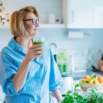 A woman in a bright kitchen holds a glass of green smoothie while standing beside fresh fruits, leafy greens, and a blender, symbolizing a natural detox or healthy cleanse.