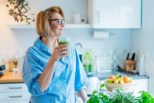 A woman in a bright kitchen holds a glass of green smoothie while standing beside fresh fruits, leafy greens, and a blender, symbolizing a natural detox or healthy cleanse.