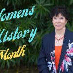 Portrait of a smiling woman standing in front of lush green foliage, wearing a dark floral blazer and orange top, with text overlay that reads “Women's Hisoty Month” in teal and gold lettering.