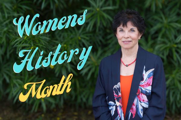 Portrait of a smiling woman standing in front of lush green foliage, wearing a dark floral blazer and orange top, with text overlay that reads “Women's Hisoty Month” in teal and gold lettering.