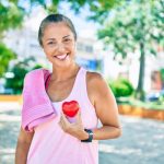Smiling woman outdoors in workout attire holding a red heart-shaped object, with a towel over her shoulder and greenery in the background.
