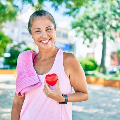 Smiling woman outdoors in workout attire holding a red heart-shaped object, with a towel over her shoulder and greenery in the background.