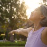 Middle-aged woman with gray hair standing outdoors in sunlight, eyes closed and arms outstretched, embracing a sense of calm, vitality, and well-being in a natural setting.