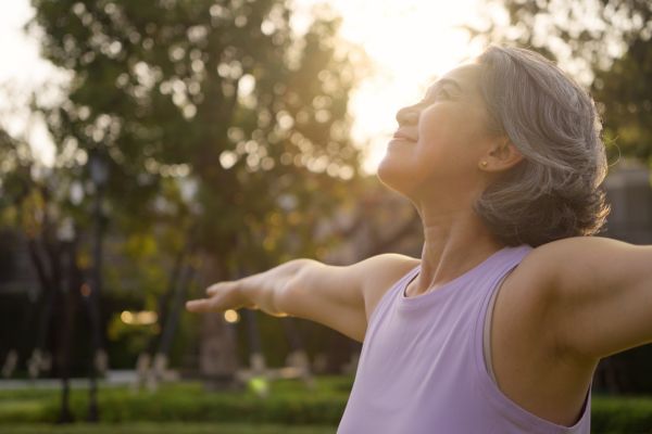 Middle-aged woman with gray hair standing outdoors in sunlight, eyes closed and arms outstretched, embracing a sense of calm, vitality, and well-being in a natural setting.