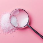 Magnifying glass focusing on white granulated sugar crystals against a soft pink background, symbolizing hidden or closely examined sugar.