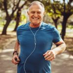 Smiling older man jogging outdoors on a sunny park trail, wearing a blue athletic shirt and earphones, surrounded by trees.