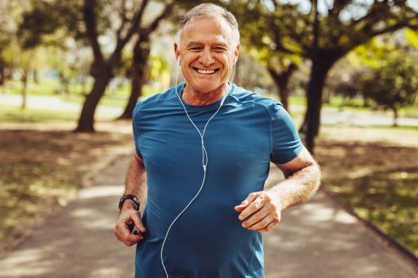 Smiling older man jogging outdoors on a sunny park trail, wearing a blue athletic shirt and earphones, surrounded by trees.