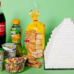 Assortment of processed foods including soda, sweetened tea, yogurt, bread, cereal, and snack bars displayed next to a large pyramid made of stacked sugar cubes on a green background, illustrating hidden sugar content.