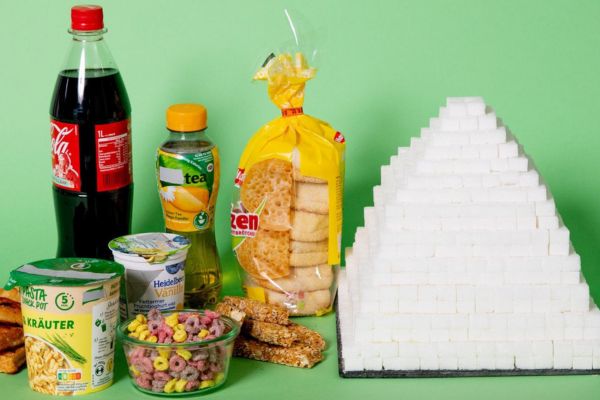 Assortment of processed foods including soda, sweetened tea, yogurt, bread, cereal, and snack bars displayed next to a large pyramid made of stacked sugar cubes on a green background, illustrating hidden sugar content.