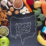 Illustration of digestive health featuring a chalk drawing of the intestines on a blackboard surrounded by whole foods including salmon, broccoli, oranges, bananas, oats, honey, seeds, nuts, garlic, carrots, bread, pasta, and fermented foods associated with gut health and nutrition.