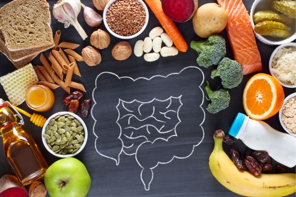 Illustration of digestive health featuring a chalk drawing of the intestines on a blackboard surrounded by whole foods including salmon, broccoli, oranges, bananas, oats, honey, seeds, nuts, garlic, carrots, bread, pasta, and fermented foods associated with gut health and nutrition.