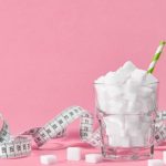 Glass filled with white sugar cubes beside a measuring tape on a pink background, symbolizing the connection between sugar consumption, weight gain, dieting, and metabolic health.