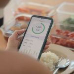 Person using a calorie-tracking app on a smartphone while eating a meal with rice, vegetables, and meal prep containers in the background.
