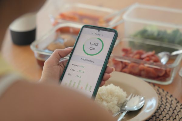 Person using a calorie-tracking app on a smartphone while eating a meal with rice, vegetables, and meal prep containers in the background.