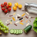 Fresh whole foods and wellness essentials arranged on a wooden table, including apples, bananas, grapes, tomatoes, cucumber, lettuce, a water bottle, and assorted supplement capsules symbolizing hydration, nutrition, and healthy living.
