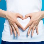 Person in a white shirt forming a heart shape with their hands over their stomach against a blue background, symbolizing gut health and digestive wellness.