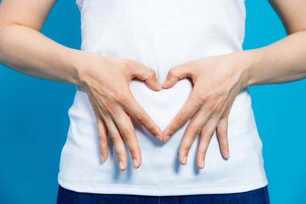 Person in a white shirt forming a heart shape with their hands over their stomach against a blue background, symbolizing gut health and digestive wellness.