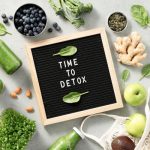 Flat lay wellness scene featuring green detox smoothies, spinach, blueberries, broccoli, ginger, apples, pumpkin seeds, and a black letter board that reads “TIME TO DETOX” on a light gray background.