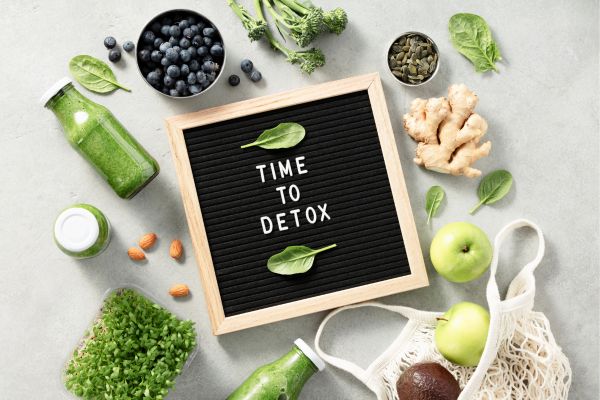 Flat lay wellness scene featuring green detox smoothies, spinach, blueberries, broccoli, ginger, apples, pumpkin seeds, and a black letter board that reads “TIME TO DETOX” on a light gray background.