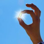 A hand holds a translucent capsule between the thumb and index finger, positioning it in front of the bright sun so it appears to glow. The clear blue sky fills the background, creating a clean, minimalist image that symbolizes supplementation, health, and wellness.