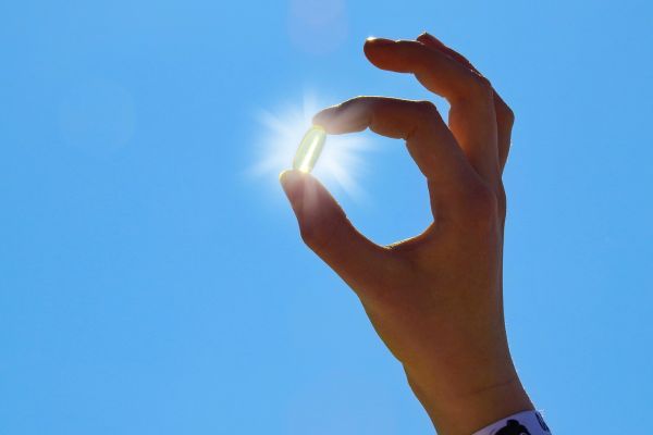 A hand holds a translucent capsule between the thumb and index finger, positioning it in front of the bright sun so it appears to glow. The clear blue sky fills the background, creating a clean, minimalist image that symbolizes supplementation, health, and wellness.