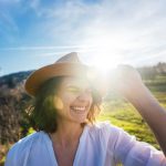A smiling woman in a sunhat enjoys a bright, sunny day outdoors in a green countryside setting, reflecting the joy and wellness benefits of spending time in nature.