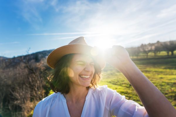 A smiling woman in a sunhat enjoys a bright, sunny day outdoors in a green countryside setting, reflecting the joy and wellness benefits of spending time in nature.