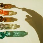 Five small glass bottles in different colors are arranged vertically on a light surface, each casting a long shadow. A hand reaches toward the top bottle, creating a dramatic silhouette across the image. Sunlight reflects through the glass, producing shimmering patterns that give the minimalist composition a clean, scientific, and wellness-focused aesthetic.