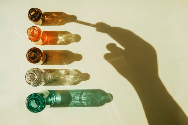 Five small glass bottles in different colors are arranged vertically on a light surface, each casting a long shadow. A hand reaches toward the top bottle, creating a dramatic silhouette across the image. Sunlight reflects through the glass, producing shimmering patterns that give the minimalist composition a clean, scientific, and wellness-focused aesthetic.