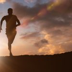 Silhouette of a person running along the crest of a hill at sunrise, with bright sunlight flaring from the horizon and dramatic orange and gray clouds filling the sky. The image conveys endurance, cardiovascular fitness, vitality, and healthy aging.