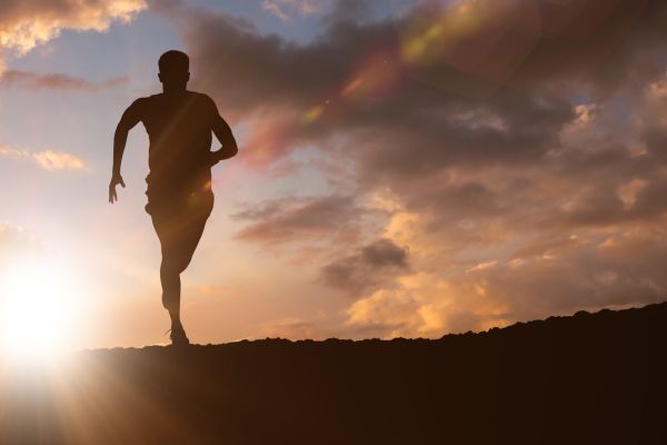 Silhouette of a person running along the crest of a hill at sunrise, with bright sunlight flaring from the horizon and dramatic orange and gray clouds filling the sky. The image conveys endurance, cardiovascular fitness, vitality, and healthy aging.