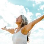 A healthy older woman stands outdoors beneath a bright blue sky with soft white clouds, eyes closed and arms stretched wide in a gesture of openness and gratitude. Sunlight illuminates her face and silver hair, creating a peaceful, uplifting scene that conveys vitality, healthy aging, freedom, and overall well-being.