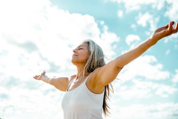 A healthy older woman stands outdoors beneath a bright blue sky with soft white clouds, eyes closed and arms stretched wide in a gesture of openness and gratitude. Sunlight illuminates her face and silver hair, creating a peaceful, uplifting scene that conveys vitality, healthy aging, freedom, and overall well-being.