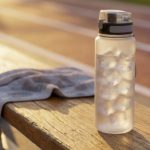 A frosted reusable water bottle filled with ice sits on a wooden bench beside a gray workout towel at an outdoor running track. Warm golden sunlight creates a bright, refreshing atmosphere, emphasizing hydration before or after exercise on a hot day.
