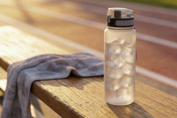 A frosted reusable water bottle filled with ice sits on a wooden bench beside a gray workout towel at an outdoor running track. Warm golden sunlight creates a bright, refreshing atmosphere, emphasizing hydration before or after exercise on a hot day.