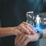 A person wearing a black shirt interacts with a smartwatch on their wrist while semi-transparent digital health icons float above the device. The holographic interface displays symbols representing medical records, heart health, lungs, stomach, and other biometric data, illustrating wearable technology used to monitor personal health and wellness. The background is softly blurred, keeping the focus on the smartwatch and futuristic health dashboard.