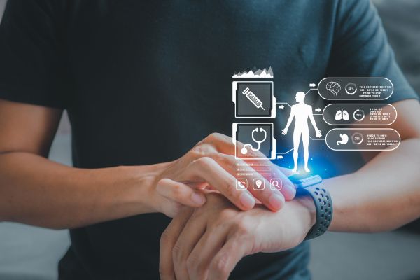 A person wearing a black shirt interacts with a smartwatch on their wrist while semi-transparent digital health icons float above the device. The holographic interface displays symbols representing medical records, heart health, lungs, stomach, and other biometric data, illustrating wearable technology used to monitor personal health and wellness. The background is softly blurred, keeping the focus on the smartwatch and futuristic health dashboard.