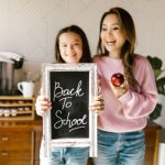 A smiling mother and daughter stand together in a kitchen, holding a chalkboard sign that reads “Back To School,” while the mother holds a red apple.