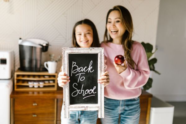 A smiling mother and daughter stand together in a kitchen, holding a chalkboard sign that reads “Back To School,” while the mother holds a red apple.