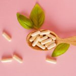 A wooden spoon filled with white supplement capsules, surrounded by green leaves and scattered capsules on a soft pink background.