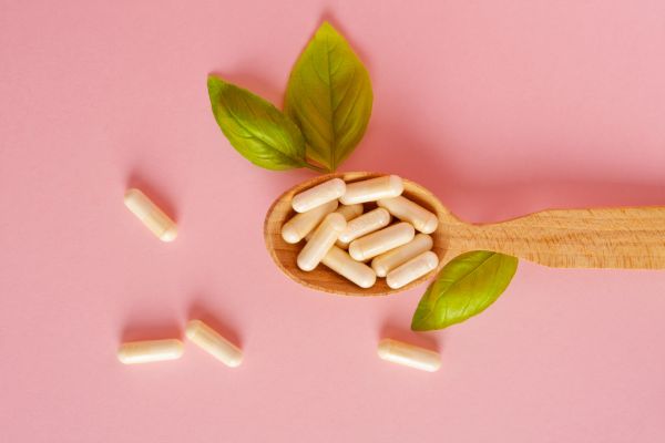 A wooden spoon filled with white supplement capsules, surrounded by green leaves and scattered capsules on a soft pink background.