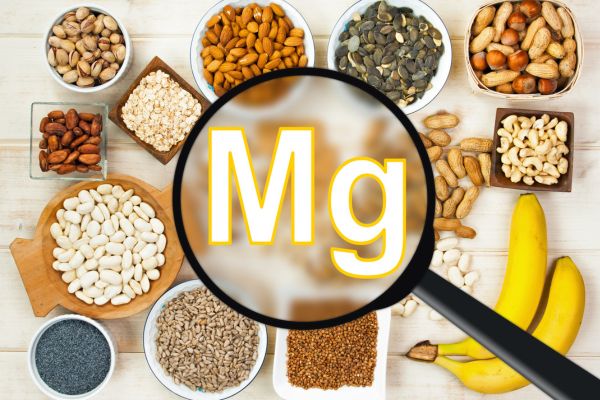 Assortment of magnesium-rich foods, including almonds, pumpkin seeds, peanuts, beans, sunflower seeds, and bananas, surrounding a magnifying glass highlighting the chemical symbol “Mg” for magnesium.