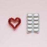 A red beaded heart beside a blister pack of white pills on a pale pink background, representing medication and heart health.