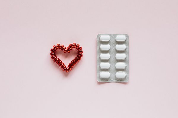 A red beaded heart beside a blister pack of white pills on a pale pink background, representing medication and heart health.