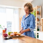 A smiling mature woman slices fresh oranges in a bright kitchen, surrounded by grapefruit, apples, raspberries, and juice.
