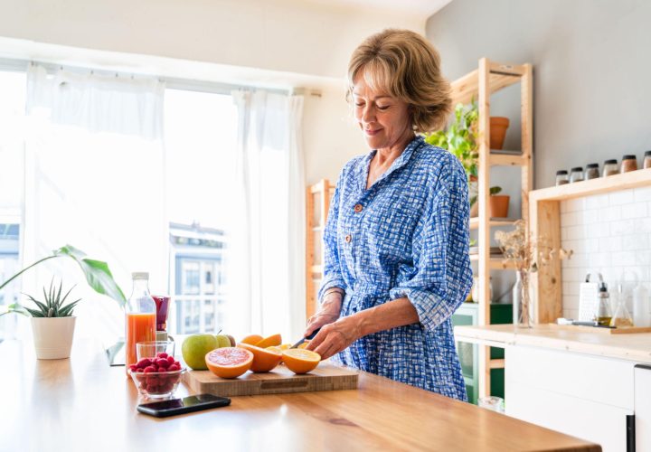 A smiling mature woman slices fresh oranges in a bright kitchen, surrounded by grapefruit, apples, raspberries, and juice.