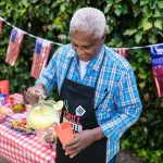 An older man wearing a “Grill Master” apron pours lemonade at an outdoor Labor Day cookout decorated with American flags and a red-checkered tablecloth.